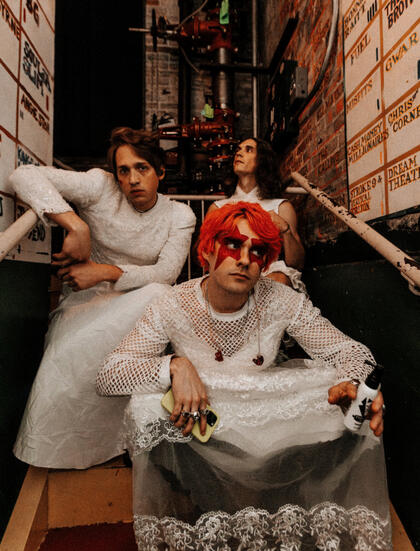 the band waterparks (left to right: guitarist geoff wigington, frontman awsten knight, drummer otto wood) seated on stairs wearing wedding dresses